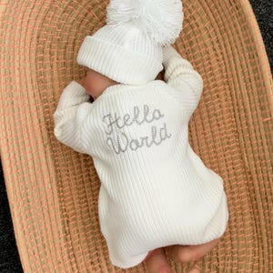May include: White knitted baby onesie with a white pom-pom hat. The onesie has the words "Hello World" embroidered on the back in silver thread.