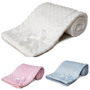 May include: A white baby blanket with a textured, dotted pattern and a satin ribbon bow. The blanket is folded and rolled, highlighting its soft texture. Additional blankets in pink and blue are also visible.