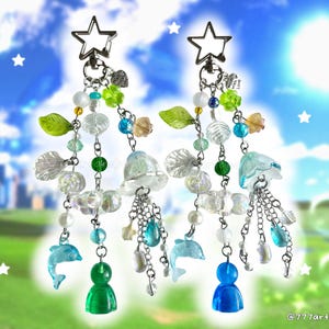 May include: Two dangling charms featuring silver star-shaped clasps, clear and colored beads, green leaf accents, small dolphin figures, and translucent bell-shaped charms. One charm features a green base bead, the other a blue base bead. Text reads 777artandjewelz.