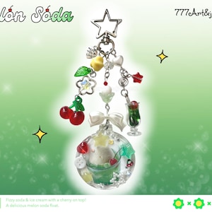 May include: A whimsical keychain featuring a clear, round charm with a melon soda float design. The charm is adorned with red cherries, a white bow, and various charms, including a star, hearts, and a miniature soda glass. The text "Melón Sóda" is at the top.