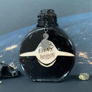 Könnte beinhalten: Eine schwarze, runde Glasflasche mit einem strukturierten, tropfenden Wachsdeckel. Die Flasche hat ein weißes Etikett mit dem Text "EVENT HORIZON" und einen silbernen Spiralanhänger. Der Hintergrund zeigt einen Sternenhimmel.