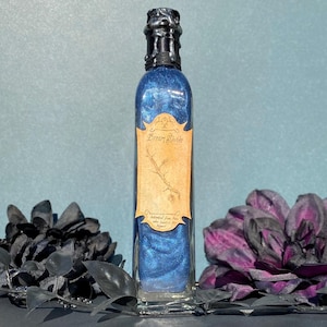 May include: A tall, rectangular glass bottle filled with shimmering blue liquid. The bottle has a dark, textured stopper and a label that reads "Dream Shade." The label features a drawing of a branch. The bottle is surrounded by dark floral accents.