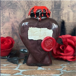 May include: A heart-shaped, burgundy-colored bottle with a red wax seal and a dripping red top. The bottle has a label with handwritten text and a decorative chain. Red roses and a brick background complete the gothic aesthetic.