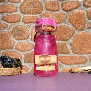 Puede incluir: Una pequeña botella de vidrio llena de un líquido rosa brillante, sellada con un corcho y cera rosa. Una etiqueta con un diseño de calavera está adherida. El fondo es una pared de piedra.