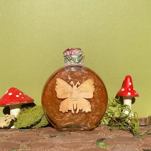 May include: A round, brown glass bottle labelled "Fairy Tonic" with a butterfly design. The bottle has a decorative stopper and is surrounded by miniature mushrooms and moss, set against a green background.