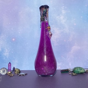 Könnte beinhalten: Eine hohe, violette Glas-Zaubertrankflasche mit dekorativem Verschluss und einem kleinen, hängenden Fläschchen. Die Flasche ist mit einer schimmernden, glitzernden Flüssigkeit gefüllt. Verschiedene Kristalle und Anhänger sind um die Basis der Flasche angeordnet.