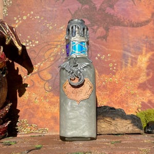 Puede incluir: Una botella de vidrio decorativa con una tapa plateada y una etiqueta que dice "Shift". La botella tiene un dije de dragón plateado y contiene un líquido brillante de color gris claro. La botella mide aproximadamente 15 cm de altura.