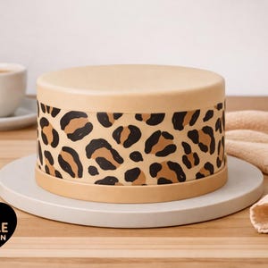 May include: A cylindrical cake covered in smooth light tan frosting with a wide horizontal band featuring a black and brown leopard print pattern. The cake sits on a round white plate, and a black heart-shaped logo with the text EDIBLE BY DESIGN .co.uk is visible in the lower left corner.