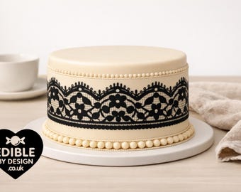 May include: A round cake with smooth cream-colored frosting, decorated with a wide band of intricate black lace-style floral patterns. The base and top edges are bordered with a row of small, uniform cream-colored beads. A black heart logo reads EDIBLE BY DESIGN .co.uk.