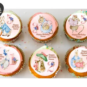 May include: Six cupcakes topped with round edible toppers featuring watercolour illustrations of Peter Rabbit characters, numbers one through six, and the text Happy Birthday Name. The toppers are printed on a light pink background with floral accents.