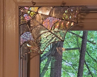 May include: A corner-shaped stained glass spider web suncatcher featuring iridescent, textured glass panels held together by thin metal solder lines. The piece is mounted in the upper corner of a window frame, displaying a reflective surface.