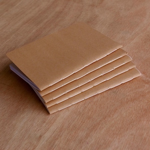 May include: A stack of five small, square notebooks with light brown covers and rough, deckled edges. The notebooks are stacked on a wooden surface with a warm, reddish-brown tone.