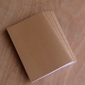 以下が含まれることがあります： ベージュ色の表紙と白い紙のページが付いた、小さな空白のノートの束。 ノートは、明るい茶色の木製の表面に斜めに積み重ねられています。 表紙は無地でニュートラルな色です。