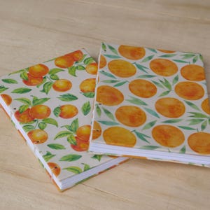 May include: Two notebooks with covers featuring watercolour illustrations of oranges and green leaves. One notebook has a pattern of oranges and leaves, while the other has a pattern of orange slices. The notebooks are on a light wood surface.