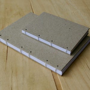 May include: Two handmade notebooks with kraft paper covers and white pages. The books are stacked, showing the stitching along the spine. They rest on a light brown wooden surface.