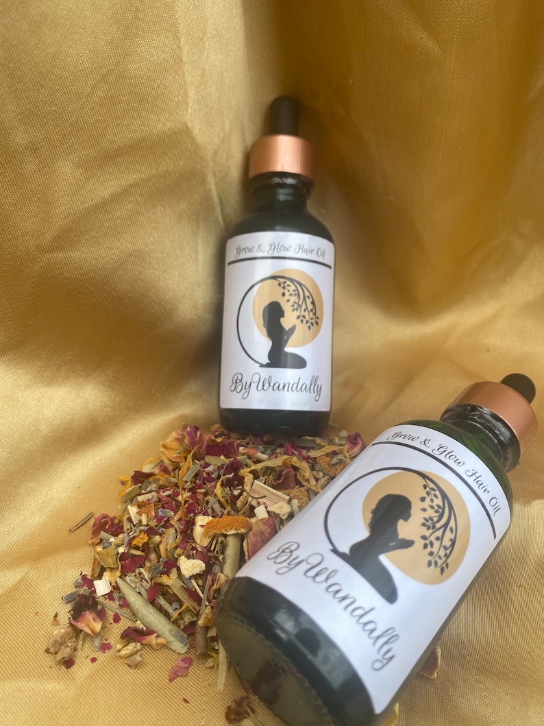 Pode incluir: Duas garrafas de vidro verde escuro com conta-gotas dourados e etiquetas brancas. As etiquetas apresentam uma silhueta de uma pessoa com um ramo de &aacute;rvore e o texto "Grow & Glow Hair Oil" e "By Wandally". As garrafas est&atilde;o sobre uma superf&iacute;cie de tecido dourado com ervas e flores secas.