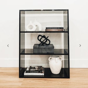 May include: A black glass shelf unit with three shelves. The shelves are filled with decorative objects, including a white ceramic vase, a black metal sculpture, a gray and white patterned box, and a white ceramic vase with two handles. The shelf unit is sitting on a wooden floor.
