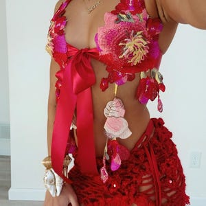 Puede incluir: Un top halter rojo con grandes bordados florales vibrantes en tonos rosa, rojo y amarillo, acentuado con un lazo de satén en el centro. La falda de ganchillo roja a juego presenta un diseño de tejido abierto y lazos laterales.