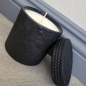 May include: Black candle in a decorative jar with a floral design. The jar has a black lid with a textured surface.