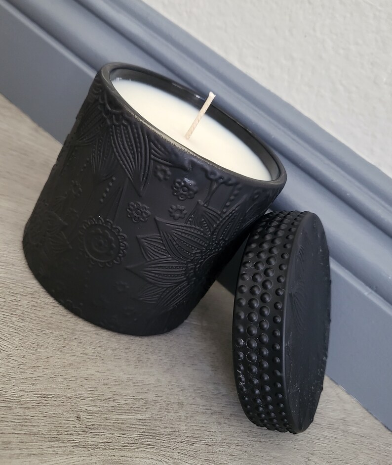 May include: Black candle in a decorative jar with a floral design. The jar has a black lid with a textured surface.