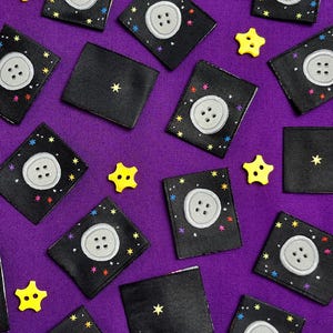 May include: A collection of black fabric patches with a white button and colourful stars on a purple background. Some patches have a single yellow star in the centre.