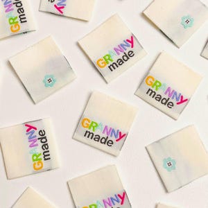 May include: A collection of white fabric squares with the text "Granny made" in colourful letters. The squares are arranged on a white surface.