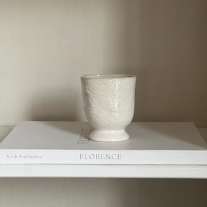 Op de afbeelding: Witte keramische vaas met een getextureerd oppervlak. De vaas staat op twee boeken met het woord "FLORENCE" op de kaft.