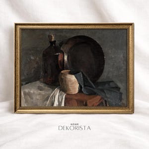 May include: A framed still life painting featuring a dark brown bottle with a cork, a round plate, and a small, light-colored cylindrical object. A dark blue cloth is draped over a table. The painting has a gold frame and the text "HOME DEKORISTA".