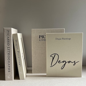 Può includere: Quattro libri con copertine in lino, impilati su una superficie bianca. I libri sono intitolati "Picasso Lithographs", "Degas Paintings", "Picasso Lithographs" e "Degas Paintings". Il libro intitolato "Degas Paintings" ha un grande "Degas" in corsivo nero sulla copertina.