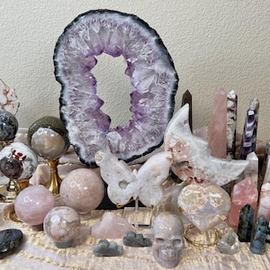 May include: A collection of various crystals and gemstones, including amethyst geodes, spheres, and polished points. The arrangement features a butterfly, a skull, and heart-shaped stones, showcasing a range of colors and textures.
