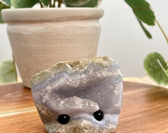 May include: A small, light blue agate geode with a natural opening, revealing a crystal-lined interior. Two black onyx beads are set into the geode, creating a whimsical face.