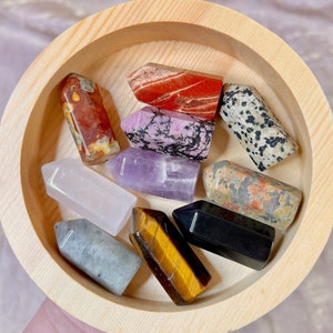 May include: A collection of ten small, pointed crystal wands in a wooden bowl. The wands are in various colors, including red, pink, purple, white, gray, black, and brown.