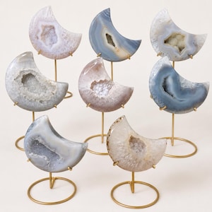 May include: Seven crescent moon-shaped agate slices with crystalline centers, each mounted on a thin gold-toned metal stand. The stones display various natural shades of gray, blue, white, and soft pink, featuring polished edges and rough, sparkling geode interiors.
