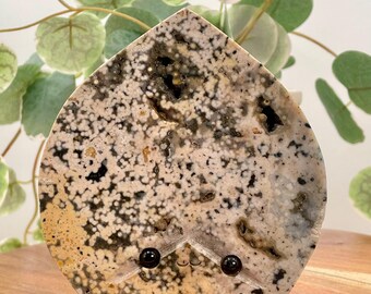 May include: A heart-shaped stone with a speckled pattern of black, white, and brown. Two black eyes are glued to the stone, giving it a whimsical look.