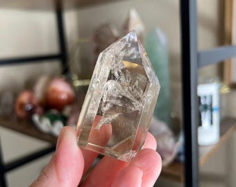 May include: A clear quartz crystal with a light brown hue, held in a hand. The crystal is faceted with a pointed top and visible internal inclusions. Other crystals and a wooden shelf are in the background.