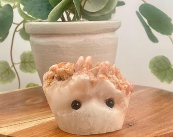 May include: A pink and orange crystal carved into the shape of a hedgehog with black eyes. The hedgehog is sitting on a wooden surface.