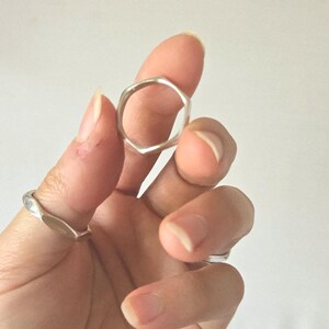 Puede incluir: Primer plano de dos anillos de plata. Uno tiene un diseño geométrico hexagonal y el otro una forma suave y curvada. Los anillos se sostienen sobre un fondo blanco liso.
