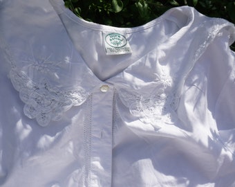 May include: A white blouse with a Peter Pan collar, adorned with lace trim and floral embroidery. The blouse has a button-down front and a label that reads "Laura Ashley". The garment is made of a soft, lightweight fabric.