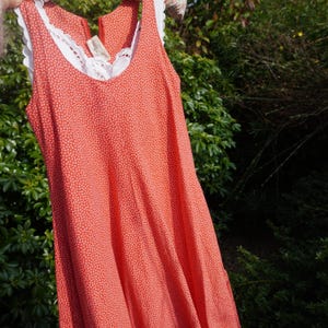 May include: A coral-coloured sleeveless dress with a white floral pattern and white lace trim around the neckline. The dress has a V-neck and a zip closure. The dress is hanging in front of a green, leafy background.