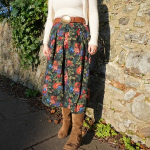 May include: A woman wearing a white sweater, a brown belt with a large gold buckle, and a long black skirt with a floral print. She is wearing brown boots.