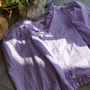 May include: A lilac button-up blouse with a delicate white floral print. The blouse features a frilled collar and cuffs, and white buttons. The sleeves are long and puffed. The garment is made from a lightweight material.