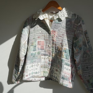 May include: A quilted jacket with a patchwork design in pastel colors, including pink, green, and blue. The jacket features a white lace collar and is buttoned up. It hangs on a wooden hanger.