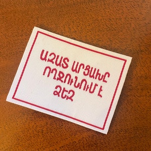 May include: White fabric patch with a red border and red embroidered text in Georgian: "შეუს უფლება არავის არ აქვს".