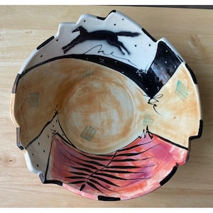 May include: A decorative ceramic bowl with an unusual, asymmetrical shape. The bowl features a black fox leaping across a white and black section, with abstract designs and a mix of colours including peach, pink, and black.