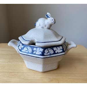 May include: A white ceramic serving dish with a blue and white floral design. The lid features a white rabbit figurine.