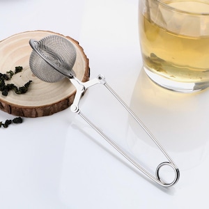 May include: A silver tea infuser with a round mesh ball and a spring-loaded handle. The infuser is resting on a wooden surface.