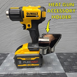 May include: A yellow and black DeWalt 20V heat gun with a silver accessory holder attached to the bottom. The holder has two nozzle attachments. The heat gun is sitting on a black and gray textured surface.