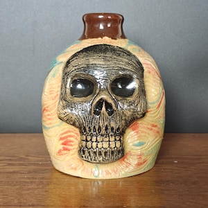 May include: A ceramic jug featuring a prominent, raised black skull relief on the front. The body of the jug displays a swirling, marbled pattern of cream, red, and teal colours. It has a smooth, dark brown glazed neck and a rounded, bulbous shape.