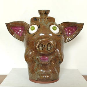 May include: A brown glazed ceramic jug shaped like a pig head with large ears, wide white eyes with green pupils, and a snout. The interior of the ears and the open mouth are painted in a glossy magenta colour, and the jug has a small spout on top.