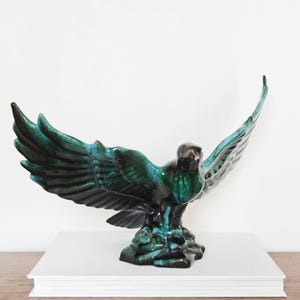 May include: A ceramic eagle figurine with a glossy finish, featuring deep emerald green and dark black glaze tones. The bird is depicted with wings fully extended in a soaring pose, perched upon a textured base that mimics a rocky surface.